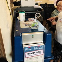 A blue drop box that has a sign on it that says DROP HERE with direction arrows and the word drop box in English and in Spanish. There's a bottle of dial hand sanitizer that is half gone sitting on top of the drop box. 