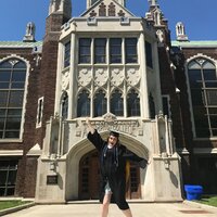 This is a picture of a woman in a graduation outfit jumping in the air in front of a school administration building. 