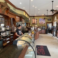 A cafe with no customers. A barista is standing at the register with a black beanie on. All of the black tables have the chairs on top of them showing you are not allowed to sit there. 