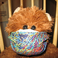 Teddy bear sitting in a wood chair wearing a multi-colored mask. 