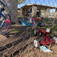 This is a picture of a discarded face mask resting next to a fence. Children's bicycles and trikes are leaned against the other side of the fence.  