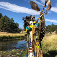 This is a picture of a Native American dressed in traditional ceremonial clothing outdoors by a stream, likely at a powwow. 