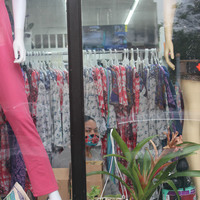 A person with a mask behind windows and plants in front of a rack of clothes.