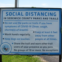 Blue and white sign by Sedgwick County, Kansas with text saying, "SOCIAL DISTANCING IN SEDGWICK COUNTY PARKS AND TRAILS: Do not use the parks or trails if you have symptoms of COVID-19 (fever, cough, shortness of breath).; Wash hands regularly.; Keep dogs on leashes.; Keep at least 6 feet away from other people at all times.; Share the trail and warn other trail users of your presence as you pass. No groups of 10 or more. No exceptions."