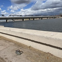 This is a picture of a batman themed face mask that has been discarded in the dirt at the edge of a lake. A rail bridge can be seen in the background. 