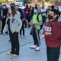 This is a picture taken of a group of people waiting in line on the street while social distancing. Every person is wearing a face mask.