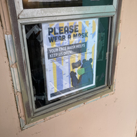 This is a picture of a sign which depicts a bear wearing a face mask and waving a flag. The sign reads: "Please wear a mask. Your face mask helps keep us open!"