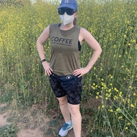 This is a picture taken of a woman outdoors, who is wearing a face mask, sun glasses, a blue Los Angeles hat, a fanny pack, a tank top which reads 'Coffee: because adulting is hard", and dark shorts. 