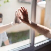 This is a picture taken of two people wearing face masks on either side of a window, and resting their hands on the same section of the window. 