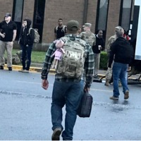 Photo of a man wearing a military backpack with a child's stuffed animal attached standing in front of a school with other men in military backpacks and full camo uniforms. 