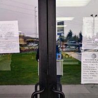 Two doors in front of a store have multiple papers taped on the inside of the glass informing patrons of their changed store hours and the decrease in people in the lobby. 