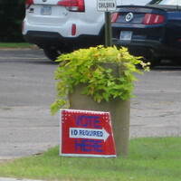 A red yard sign reading "VOTE HERE. ID required".