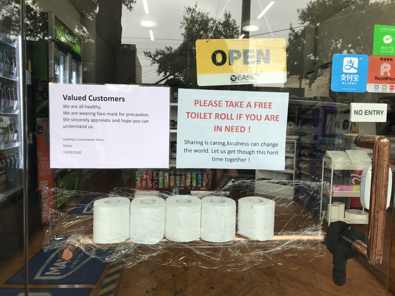 In front of a convenience store that has three signs that say: Open. Valued Customers, we are all healthy, we are wearing face masks for precautions. We sincerely appreciate and hope you can understand us. PLEASE TAKE A FREE TOILET ROLL IF YOU ARE IN NEED! Sharing is caring, kindness can change the world. Let us get through this hard time together! Below the signs is five rolls of toilet paper all in a row on top of saran wrap. 